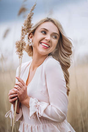 Photo of a pretty smiling girl with long blond curly hair in light long drees standing in a reed field and holding high reed branches in her handsの写真素材