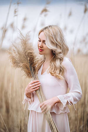 Photo of a pretty smiling girl with long blond curly hair in light long drees standing in a reed field and holding high reed branches in her handsの写真素材