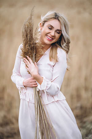 Photo of a pretty smiling girl with long blond curly hair in light long drees standing in a reed field and holding high reed branches in her handsの写真素材
