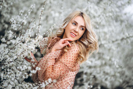Photo of a pretty smiling girl with long blond curly hair in light long drees standing in a reed field and holding high reed branches in her handsの写真素材