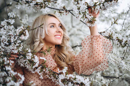 Photo of a pretty smiling girl with long blond curly hair in light long drees standing in a reed field and holding high reed branches in her handsの写真素材