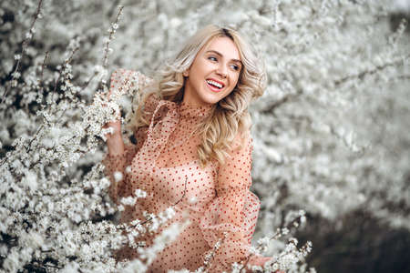 Photo of a pretty smiling girl with long blond curly hair in light long drees standing in a reed field and holding high reed branches in her handsの写真素材