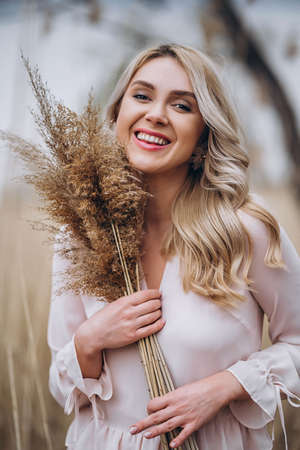 Photo of a pretty smiling girl with long blond curly hair in light long drees standing in a reed field and holding high reed branches in her handsの写真素材