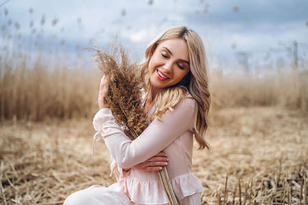 Photo of a pretty smiling girl with long blond curly hair in light long dress standing in a reed field and holding high reed branches in her handsの写真素材