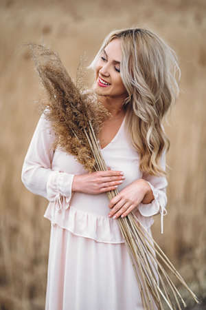 Photo of a pretty smiling girl with long blond curly hair in light long drees standing in a reed field and holding high reed branches in her handsの写真素材