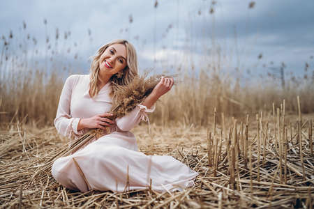 Photo of a pretty smiling girl with long blond curly hair in light long dress standing in a reed field and holding high reed branches in her handsの写真素材