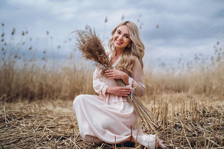 Photo of a pretty smiling girl with long blond curly hair in light long drees standing in a reed field and holding high reed branches in her handsの写真素材