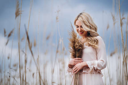 Photo of a pretty smiling girl with long blond curly hair in light long drees standing in a reed field and holding high reed branches in her handsの写真素材