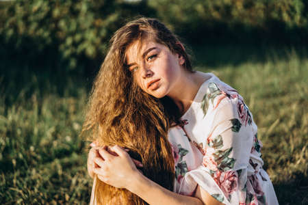 Full length portrait of beautiful woman with long curly hair on currant field background. Girl in a light dress sits on the grass in sunny day.の写真素材