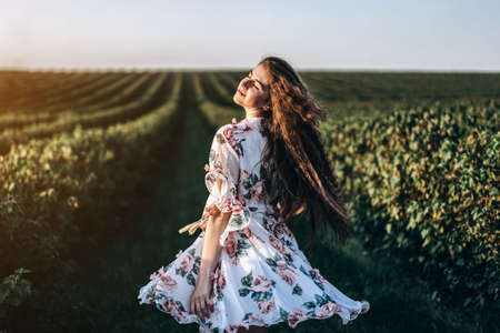 Full length portrait of beautiful woman with long curly hair on currant field background. Girl in a light dress sits on the grass in sunny day.の写真素材