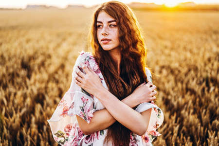 Full length portrait of beautiful woman with long curly hair on currant field background. Girl in a light dress sits on the grass in sunny day.の写真素材
