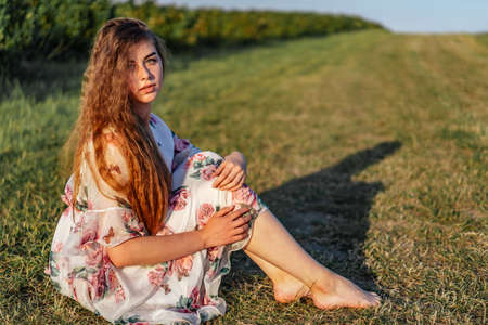 Full length portrait of beautiful woman with long curly hair on currant field background. Girl in a light dress sits on the grass in sunny day.の写真素材
