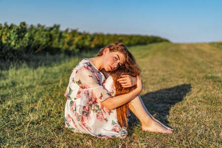 Full length portrait of beautiful woman with long curly hair on currant field background. Girl in a light dress sits on the grass in sunny day.の写真素材