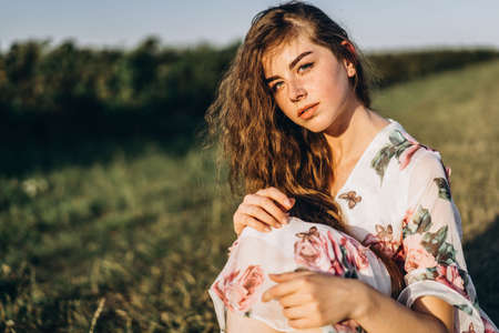 Full length portrait of beautiful woman with long curly hair on currant field background. Girl in a light dress sits on the grass in sunny day.の写真素材