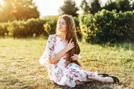 Full length portrait of beautiful woman with long curly hair on currant field background. Girl in a light dress sits on the grass in sunny day.の写真素材