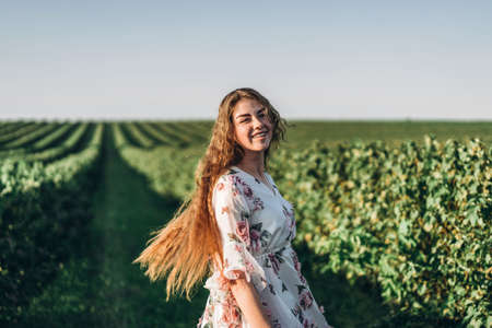 Full length portrait of beautiful woman with long curly hair on currant field background. Girl in a light dress sits on the grass in sunny day.の写真素材