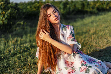 Full length portrait of beautiful woman with long curly hair on currant field background. Girl in a light dress sits on the grass in sunny day.の写真素材