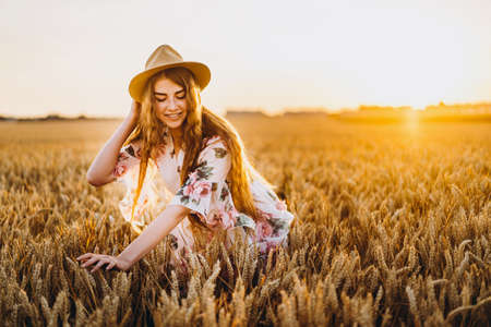 Full length portrait of beautiful woman with long curly hair on currant field background. Girl in a light dress sits on the grass in sunny day.の写真素材