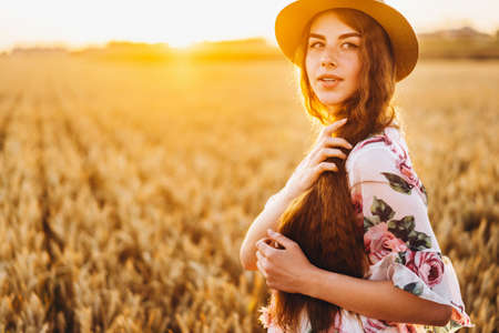 Full length portrait of beautiful woman with long curly hair on currant field background. Girl in a light dress sits on the grass in sunny day.の写真素材