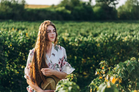 Full length portrait of beautiful woman with long curly hair on currant field background. Girl in a light dress sits on the grass in sunny day.の写真素材