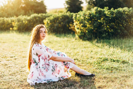 Full length portrait of beautiful woman with long curly hair on currant field background. Girl in a light dress sits on the grass in sunny day.の写真素材