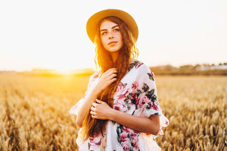 Full length portrait of beautiful woman with long curly hair on currant field background. Girl in a light dress sits on the grass in sunny day.の写真素材