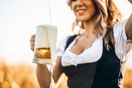 Close-up of the girls hands in dirndl, traditional festival dress, pouring a full glass of beer with huge foam, outdoors with blurred background. Oktoberfest, St. Patrickâs day, international beer day conceptの写真素材