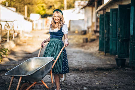 Pretty smiling blonde in traditional dress with wheelbarrow working in backyard of the farmの写真素材