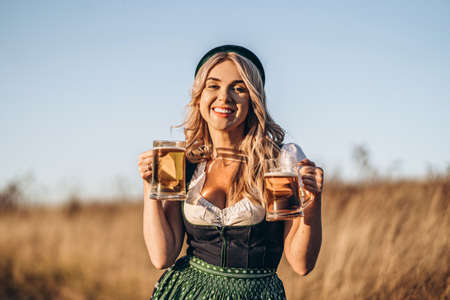 Pretty happy blonde in dirndl, traditional festival dress, holding two mugs of beer outdoors in the field with blurred background. Oktoberfest, St. Patrickâs day, international beer day concept.の写真素材