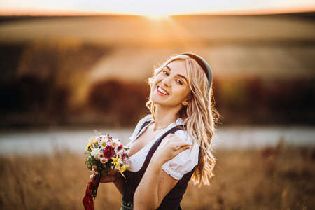 Pretty blonde girl in dirndl, traditional festival dress, standing outdoors in the field at sunset time, holding bouquet of a field flowers. Oktoberfest, St. Patrickâs day, international beer day conceptの写真素材