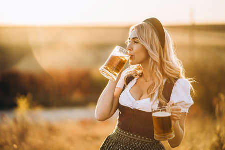 Pretty happy blonde in dirndl, traditional festival dress, holding two mugs of beer outdoors in the field with blurred background. Oktoberfest, St. Patrickâs day, international beer day concept.の写真素材