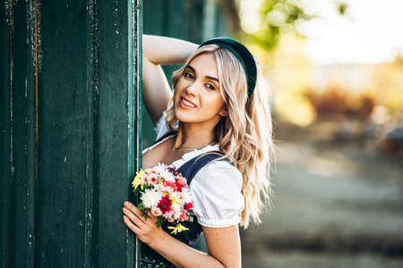 Pretty blonde in dirndl, traditional oktoberfest dress and headband, with bouquet of field flowers in hands standing on the farm near wooden doorの写真素材
