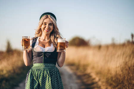 Pretty happy blonde in dirndl, traditional festival dress, holding two mugs of beer outdoors in the field with blurred background. Oktoberfest, St. Patrickâs day, international beer day concept.の写真素材