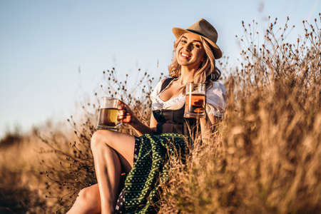 Pretty happy blonde in dirndl, traditional festival dress, sitting with two mugs of beer outdoors in the field with blurred background. Oktoberfest, St. Patrickâs day, international beer day concept.の写真素材