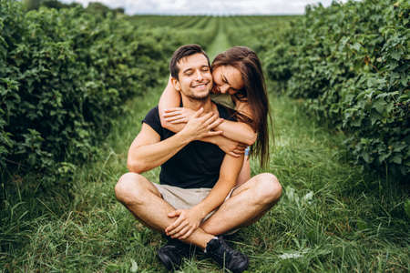 A man sitting on green grass between currant bushes, a woman with long hair hugging him from behind. Background of currant plantations.の写真素材