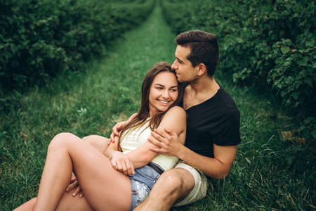 Woman with long hair and man sitting between currant bushes, hugging and kissing. Background of green currant plantations.の写真素材