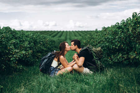 Man and woman with backpacks on their backs sitting hugging on the green grass between the currant bushes. Walking on the currant plantations in the countryside.の写真素材