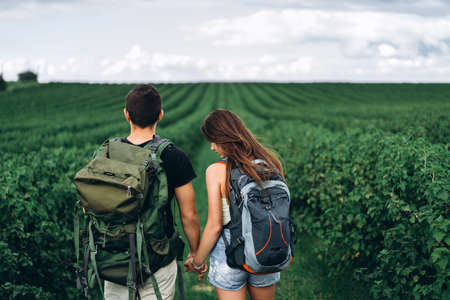 Back view of young couple with backpacks on currant plantations holding hands and spending time together. Love, tourism concept.の写真素材