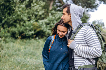 Young couple with backpacks on their backs in the forest. Loving man kisses his beautiful girlfriend on a hike in the woods.の写真素材