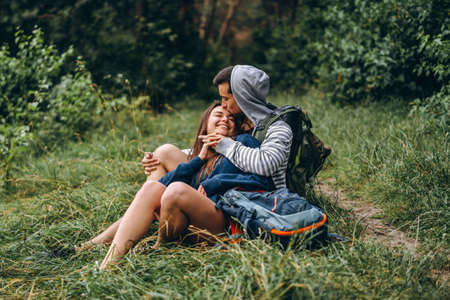 Woman with long hair and man sitting on the grass in the forest with backpacks. Have fun in nature, hugging and kissing.の写真素材
