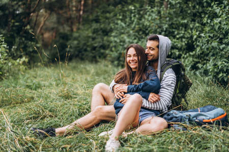 Woman with long hair and man sitting on the grass in the forest with backpacks. Have fun in nature, hugging and kissing.の写真素材