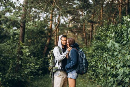 Young couple with backpacks on their backs in the forest. Loving man kisses his beautiful girlfriend on a hike in the woods.の写真素材
