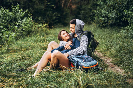 Woman with long hair and man sitting on the grass in the forest with backpacks. Have fun in nature, hugging and kissing.の写真素材
