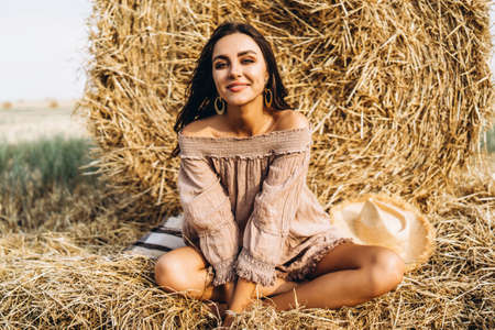 A young woman with long hair and in a dress sits near a hay bale. Woman posing smiling and looking at camera.の写真素材