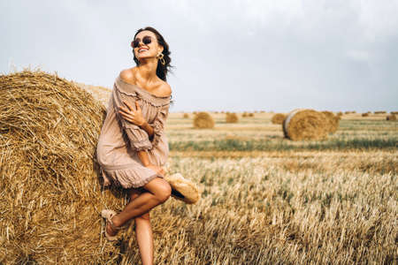 Beautiful young girl with long hair in sunnglasses and straw hat posing on a wheat field near hay bales. Happy brunette in summer dress.の写真素材