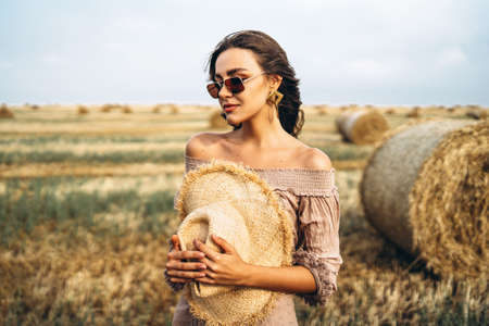 Smiling woman in sunglasses with bare shoulders on a background of wheat field and bales of hay.の写真素材