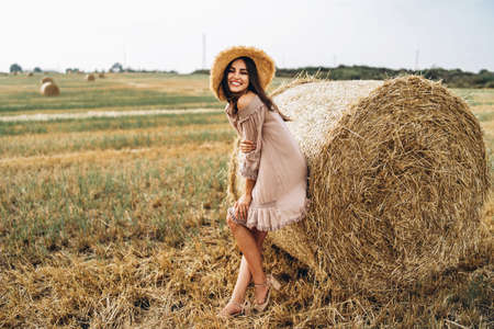 Beautiful young girl with long hair in sunnglasses and straw hat posing on a wheat field near hay bales. Happy brunette in summer dress.の写真素材