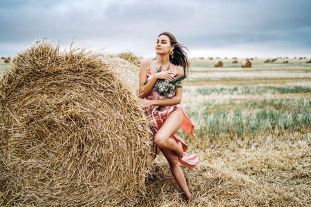 Young brunette in a pink satin dress, without linen standing near hay bales in windy weather. A woman holds a bouquet of wildflowers.の写真素材