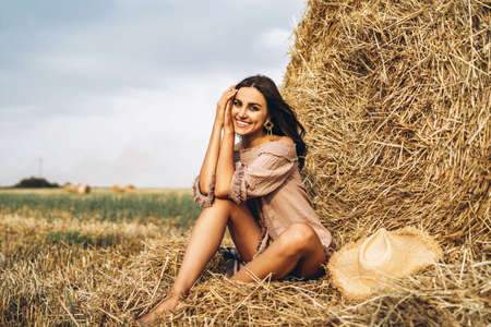 A young woman with long hair and in a dress sits near a hay bale. Woman posing smiling and looking at camera.の写真素材
