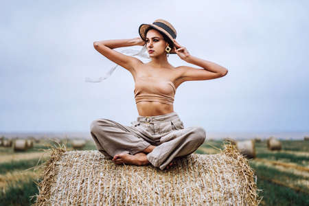 Barefoot brunette in linen pants and bare shoulders sitting on a hay bales in warm autumn day. Behind her is a wheat field.の写真素材